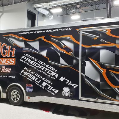 A snowmobile trailer with a custom vinyl wrap with black, orange, and white tears showing checkered flag and logos.