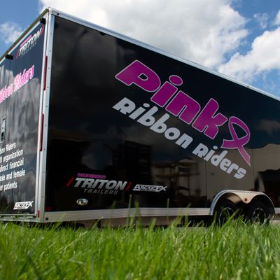 A snowmobile trailer with a custom vinyl wrap with pink ribbon riders logo and white text.