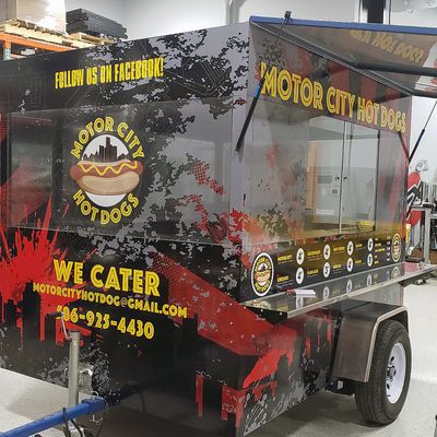 A commercial food hotdog truck with a custom vinyl wrap with yellow, red, and gray logos and text with a grunge city design.
