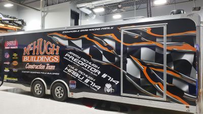 A snowmobile trailer with a custom vinyl wrap with black, orange, and white tears showing checkered flag and logos.