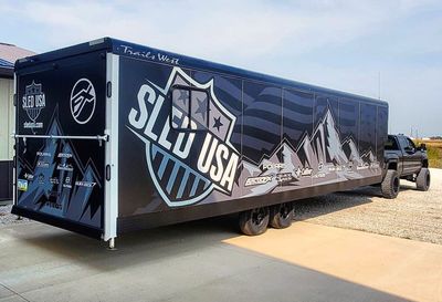 A snowmobile trailer with a custom vinyl wrap with black, gray, and white mountains and flags.